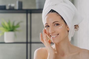 Smiling latina female in towel moisturizes facial skin with cream in bathroom. Skincare treatment
