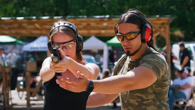 Outdoor closeup shot of focused caucasian skinny woman in protective headphones and glasses aiming at the target. Muscular shooting instructor correcting her posture. High quality 4k footage