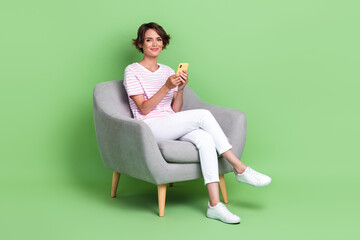 Fototapeta na wymiar Full size photo of cute young woman sit armchair relaxing hold device typing dressed trendy striped look isolated on green color background