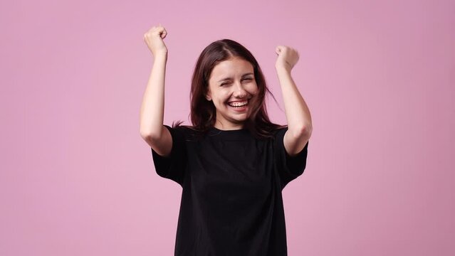 4k video of positive woman in a balck T-shirt with raised arms and a smile on pink background.
