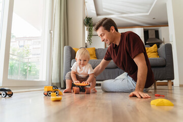 Cute blond baby boy sitting on orange skateboard on floor in living-room playing with daddy in...