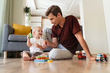 Obraz na płótnie Canvas Cute adorable baby boy with blond hair playing sorter sitting on floor with his father in living-room, spending time together after napping. Caring daddy. Dad playing with his son using education toys