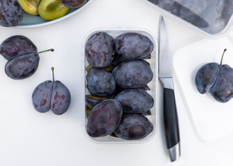 Whole and halves of a blue plums lie on a plate and in plastic box. Autumn harvest. Preparatory stage for making jam, home baking or freezing for the winter.
