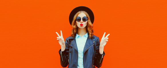 Portrait of stylish woman model blowing her red lips sending air kiss wearing black rock style leather jacket, round hat on red background