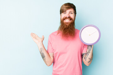 Young caucasian man holding a clock isolated on blue background showing a copy space on a palm and holding another hand on waist.