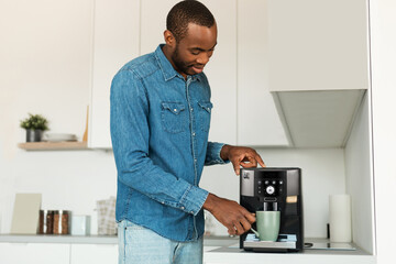 Handsome young african american man using modern coffee machine in kitchen, making fresh aromatic...