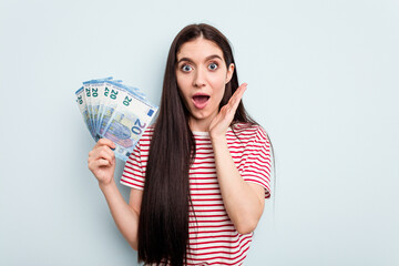 Young caucasian woman holding banknotes isolated on blue background surprised and shocked.