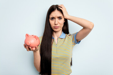 Young caucasian woman holding a piggy bank isolated on blue background being shocked, she has...