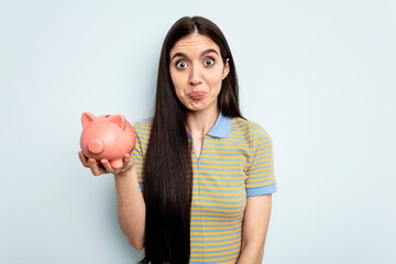 Young caucasian woman holding a piggy bank isolated on blue background shrugs shoulders and open eyes confused.