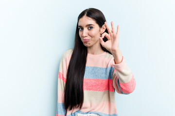 Young caucasian woman isolated on blue background winks an eye and holds an okay gesture with hand.