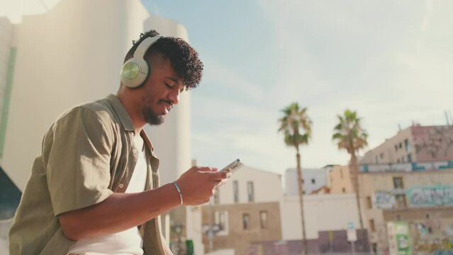 Close up, young male student dressed in an olive color shirt sits outside next to the university, listens to music on headphones, selects tracks on his phone. Camera moves backwards  