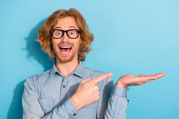 Portrait of astonished excited handsome guy indicate finger hand hold empty space isolated on blue...