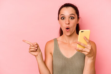 Young caucasian woman holding mobile phone isolated on pink background pointing to the side