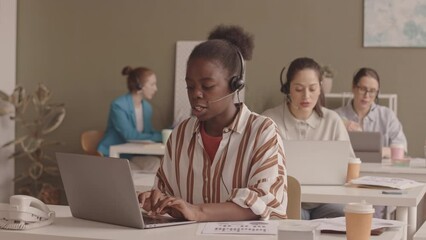 Waist up slowmo of young African American woman and her female colleagues working at call center in office, speaking to clients through headsets and entering data on laptops - Powered by Adobe