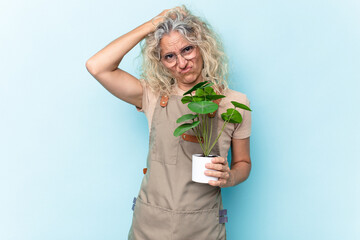 Middle age gardener woman holding a plant isolated on blue background being shocked, she has remembered important meeting.