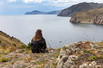 Fototapeta premium A tourist girl sits on a high rock and admires a beautiful view of the shore of Baikal Lake. Journey along the lake, outdoor recreation