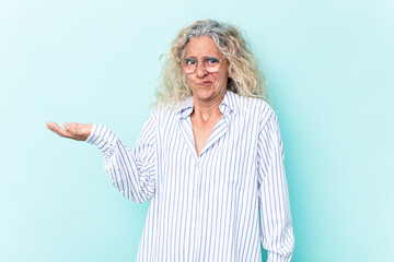 Middle age caucasian woman isolated on blue background doubting and shrugging shoulders in questioning gesture.