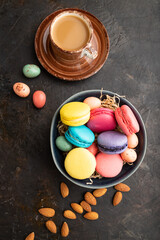 Different colors macaroons and chocolate eggs in ceramic bowl, cup of coffee on black concrete background. top view, close up.