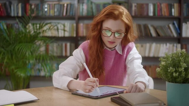 Student Woman with Red Hair holding stylus browsing on digital tablet. Freelancer interacting with tablet-pc display using touch pen. Programmer with glasses in home studio casually using touchscreen