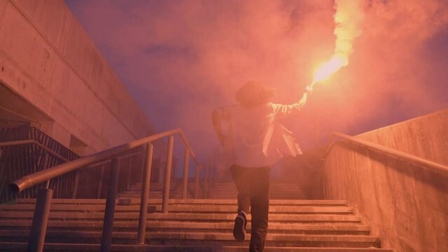 Teenager Holding Burning Flare In Raised Hand, Running On Stairs At Night, Slow Motion, Back View Tracking Shot. Teen Protest, Rebellion Demonstration, Youth, Revolution Concept