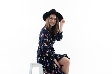 A brunette in a long floral dress with a hat poses for the camera in the studio