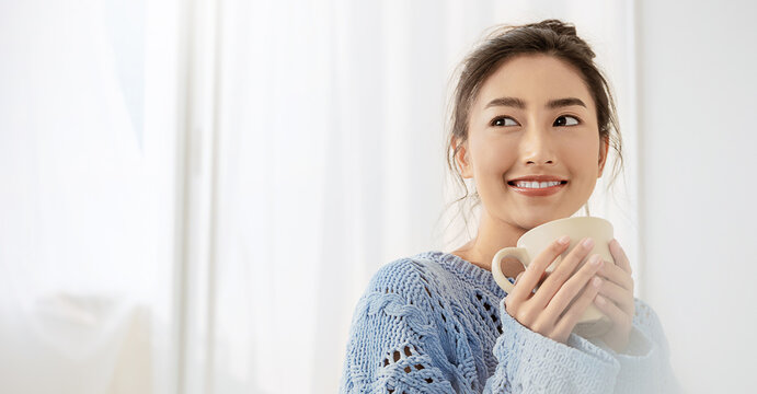 Portrait of young beautiful asian woman wear pink sweater hands holding coffee cup morning cold winter time in white bedroom. Happy cheerful relaxing in cold winter. Korean makeup lifestyle concept