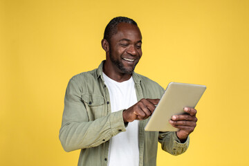 Cheerful handsome adult black man in casual typing on tablet, reading blog, isolated on yellow...