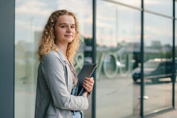 Fototapeta na wymiar Caucasian girl student holding notebook folders in her hands strolling near glass building looking at camera. Handsome woman waiting for someone. Breathing