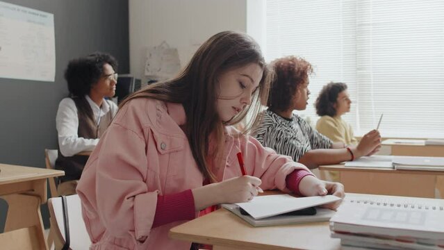 Chest-up side view of teenage Caucasian girl cheating on school test, hiding smartphone between notebook pages