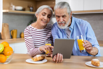 Cheerful Elderly Couple Having Breakfast Together And Using Digital Tablet In Kitchen