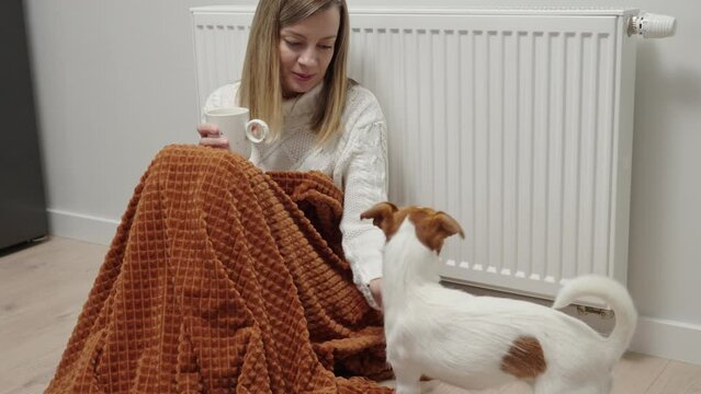 Worried sad woman and her dog sits under blanket near heating radiator and use smartphone, Rising costs in private households for gas bill due to inflation and war, Energy crisis