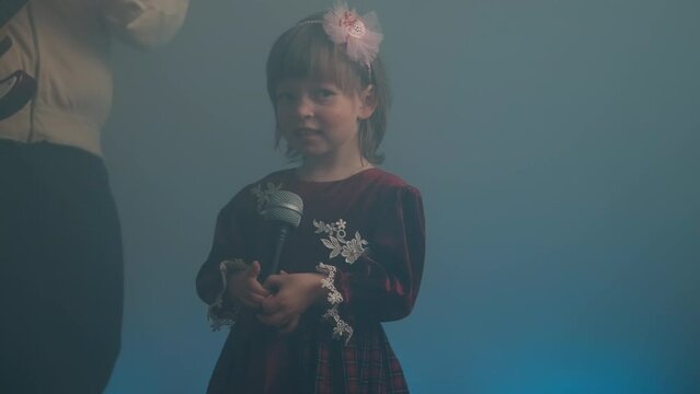 Little girl on stage in vintage dress, she sings into microphone and dances, her father plays an electric guitar. Color music is shining and smoke is billowing. Family time, creativity and hobbies