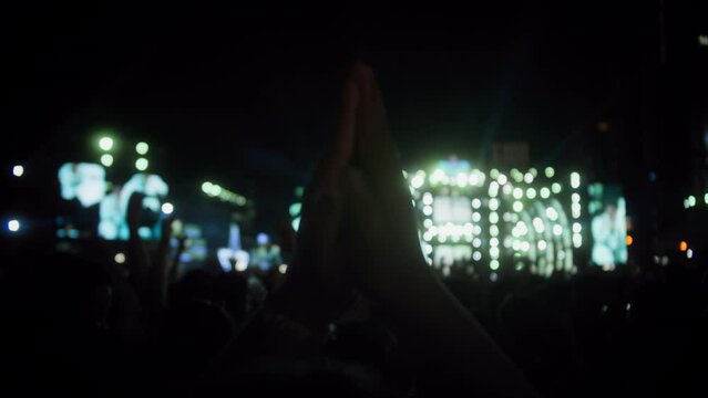 Fans raise their hands in front bright colorful strobe lights on stage. Rock band with guitarists and drummer at concert in street at night.