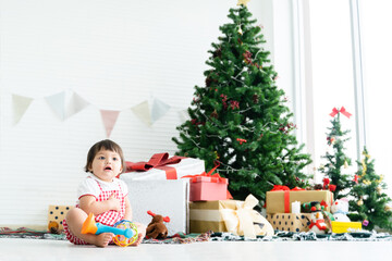 Adorable 8 months Asian baby girl smiling and having fun playing toy, sitting near a Christmas tree on floor, among gift boxes, dolls and toys at home for celebrate holiday