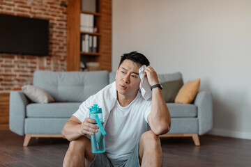After training. Tired asian man with white towel on neck holding shaker with water and swiping...
