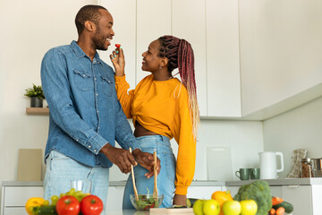 Happy black woman feeding her husband, couple preparing healthy lunch in light kitchen interior, free space