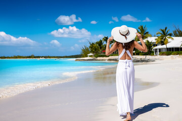 A beautiful woman in a white summer dress walks barefoot down a tropical paradise beach in the...