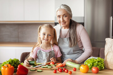 Happy little girl and old woman in aprons cut organic vegetables for salad in minimalist kitchen interior