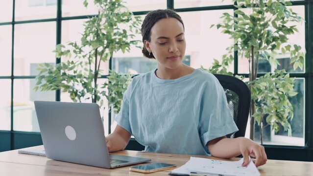 Positive smart brazilian or hispanic young businesswoman, secretary, financial manager, sitting at a workdesk in a modern office, doing paperwork, studying financial charts, analyzing profit, smiling