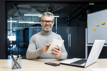 Naklejka na ściany i meble Portrait of successful programmer team leader, mature gray-haired engineer testing new app on tablet computer, businessman working inside modern office building, smiling and looking at camera