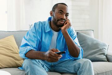 Bored black man watching boring TV, switching channels holding remote controller, sitting on sofa at home