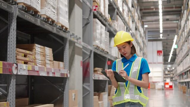 Beautiful asian engineer woman wearing safety helmet and reflective vest, She checking goods and supplies on shelves with tablet in ware house. Logistic and business export concept.
