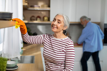 Portrait Of Smiling Senior Woman Cleaning Shelves In Kitchen From Dust