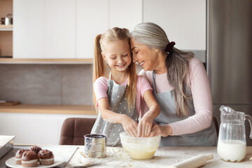 Satisfied old female in apron hugs little granddaughter, make dough, have fun in minimalist kitchen interior