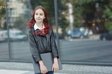 Young girl with flowing long red hair standing with a laptop at the city street in front of glass doors and looking into the distance