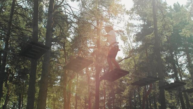 Brave teenage girl in gear and helmet, overcoming obstacles in a rope park in a sunny wooded area. Side view from below