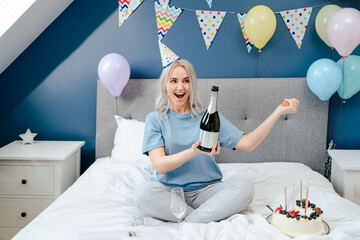 Happy emotional woman in pajama and party cap opening a bottle of champagne, sitting on the bed...