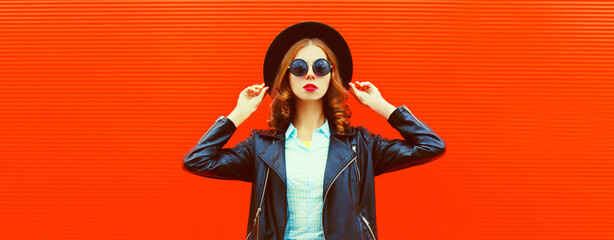 Portrait of stylish woman model posing wearing black rock style leather jacket, round hat on red background