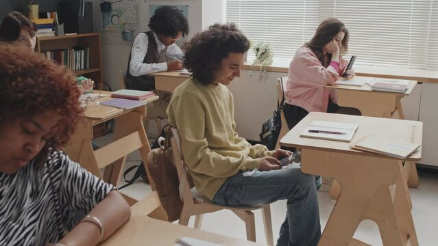 Medium long side-view of curly-haired teenage guy sitting at desk in class at daytime, receiving text message, hiding smartphone, smiling and replying