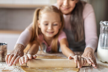 Smiling european granddaughter and elderly grandma in aprons make dough with rolling pin for pizza...